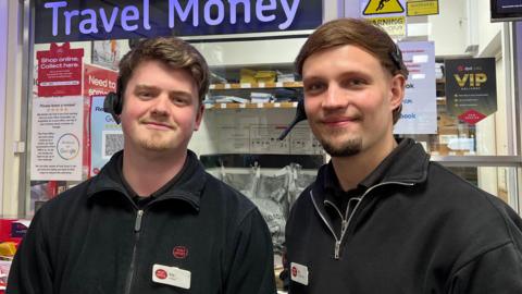 Two young men in Post Office uniforms - black fleeces and shirts - with white name badges. Behind them are bags of post and lots of notices with the Post Office's red branding.