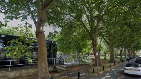 A row of tall trees are in front of a single storey glass fronted building. A covered bike rack is next to it. In front of the trees is a short metal fence, a path and a road with parked cars. 