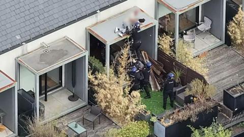 Police officers arresting a man who is on a roof. There are gardens on either side of the one that has several police officers in.