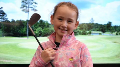 Hope in a pink zip-up sweatshirt. She is looking at the camera and is smiling while holding a golf club. She is stood in front of a screen that has a golf range on it.