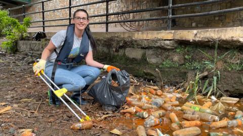 A woman wearing glasses, a grey T-shirt and blue jeans, with a life jacket over the top, crouches with a litter picker, next to piles of dirty plastic bottles.