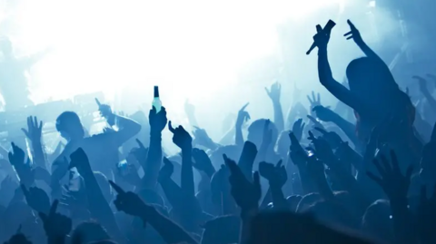 An image from behind of people raising their hands in the air, in silhouette, as a DJ stands on a stage. The image is bleached-out white and blue due to stage lighting.