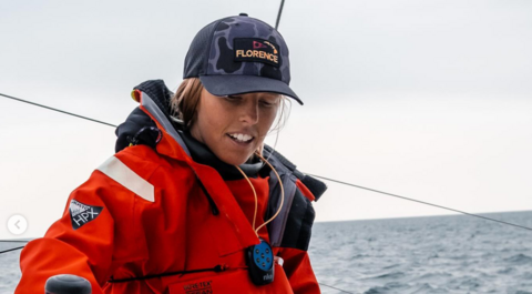 A woman in an orange sailing jacket seen with the with the sea behind her. She is also wearing a blue-camouflage baseball cap with the word "Florence" on the front.