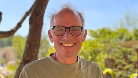 Tom Hoblyn wearing an olive green T-shirt, black glasses and smiling. He is at the Chelsea Flower Show and there is a tree trunk and bush behind him.