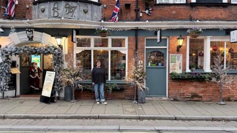 A woman standing on the pavement outside a pub that is covered in Christmas decorations. Santa is in the doorway. She is wearing jeans and a dark jumper.