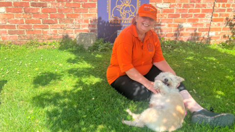 Sammy Cullis sits on the grass with a small white dog and wears an orange Food Fairies-branded polo top and hat. She is in front of a brick wall that bears the charity's logo