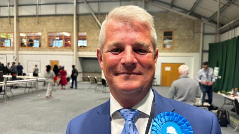 A man in a blue blazer, blue tie, white shirt with a blue rosette stands in a hall looking at the camera. 