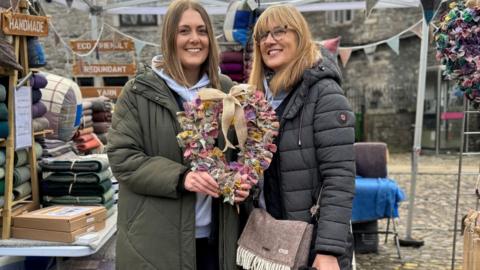 Two women stand in a market place holding a rag wreath in the shape of a heart.