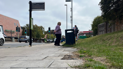 People standing at a bus stop where a bus shelter has been removed