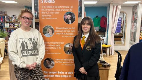 A woman and a teenager stand either side of an orange information board in a charity shop.