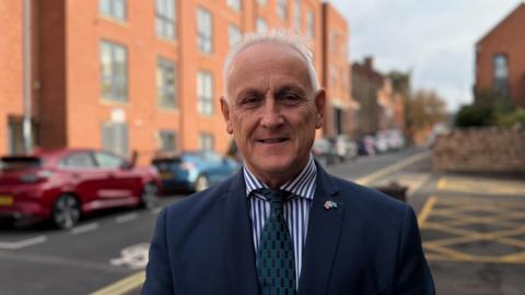 Alan Graves on a city street wearing a blue suit jacket, blue striped shirt and blue & green tie. His lapel pin features Union and Derbyshire flags crossed.