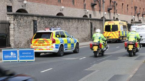 A police car and van and two motorbikes are driving along a road in Liverpool, with two blue signs nearby saying 'Project Servator' and 'vehicle checkpoint'.