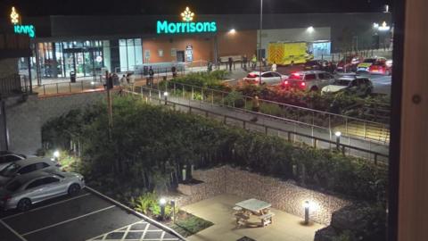 An ambulance and police cars parked outside a Morrisons supermarket along with several other vehicles in the car park. Several people are stood outside the store. It is night time and the supermarket's logo is lit up.