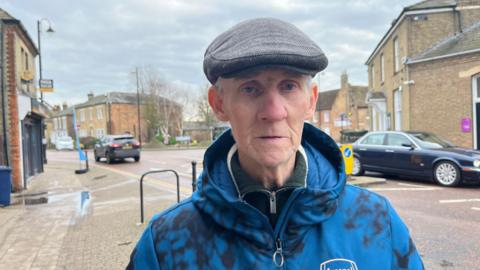 Geoffrey Taylor looking at the camera with a neutral expression. He is wearing a grey flat cap and a blue hoodie. Behind him is a pavement, part of a road and buildings.
