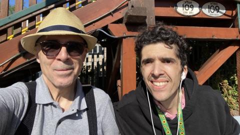 An elderly man in a hat and sunglasses next to a younger man wearing earphones outdoors in front of a building