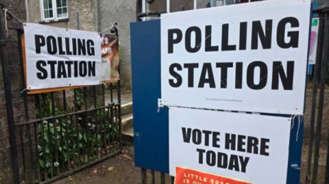 "Polling station" and "vote here today" signs, tied to railings in Devon.