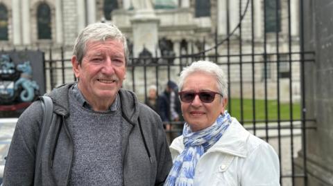 A man and a woman standing outside railings, behind them a grand looking white washed Belfast City Hall.
The man has grey hair swept to the side and blue eyes. He is smiling at the camera. He is wearing a grey hoodie over a grey jumper.
The woman is a head or so smaller than him and has white short hair. She is wearing a white coat with a white and blue scarf on. 