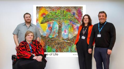 Four people are either side of the large colourful artwork hanging from a wall.