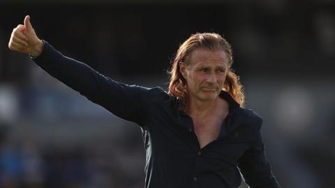Gillingham manager Gareth Ainsworth walking across pitch with right thumb held up to the fans, wearing a blue long-sleeve shirt unbuttoned to his chest