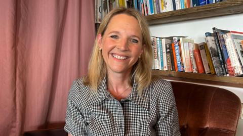 Kate has blonde hair and is wearing a black and white checked shirt. She is sat in front of a bookcase