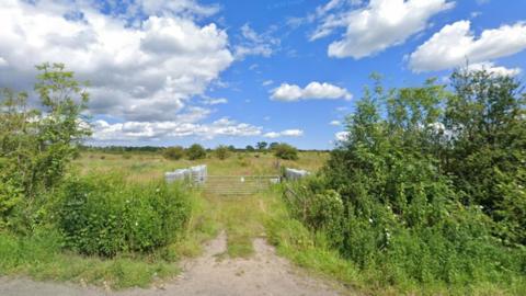 A large steel gate leads into a field in a rural location. It is summer time and the trees are full of leaves. The field beyond the gate has several trees in it and the grass is quite long. 