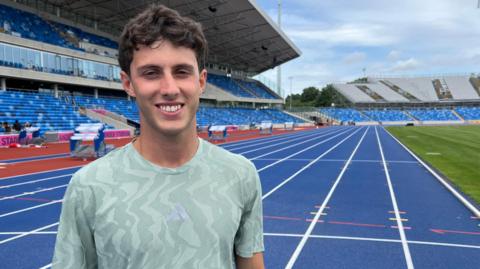 Jake Minshull stands in a light green training top on a blue race track. The white lane lines stretch into the background. The stands are behind him and the grass infield on the right 