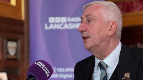 Sir Lindsay Hoyle is speaking into a microphone with the words "BBC Radio Lancashire" written on it and in the background is a large purple banner with the same words on it. 