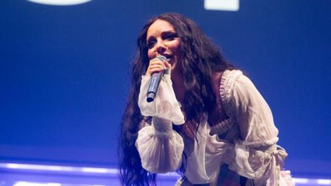 Jade Thirwall performing at the Big Weekend. She has long dark hair and is wearing a white dress. 