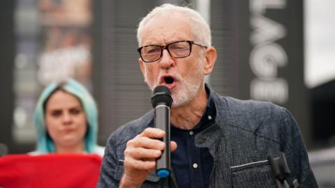 Jeremy Corbyn, a man with short grey hair, glasses and a grey beard, speaks into a microphone, held in his right hand. He is standing outside, with a woman holding a red sign and a large building in the background. He is wearing a dark jacket and shirt. 