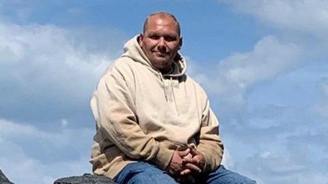 Charlie O'Sullivan in a cream coloured hoody and blue jeans sitting down on some rocks. He is looking directly at the camera against the backdrop of broken clouds.