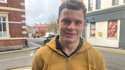 Daithí Sands. He wears a mustard hoodie and has curly brown hair. He is in his 20s