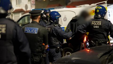 Two British Transport Police officers lead a robbery suspect into the back of a police van, they are wearing helmets and body armer. Two other police officers follow them, with another stood by the van.