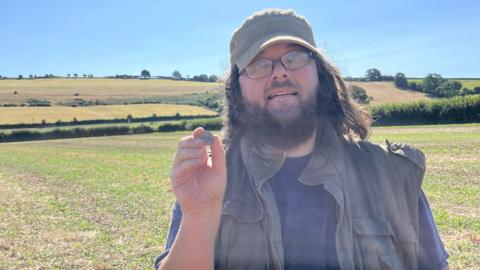 A bearded, long-haired man with glasses, wearing a green cap and body warmer holds an ancient coin a he stands in a field on a sunny day.