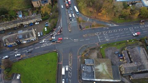 Aerial view of a busy road intersection in an urban area. Multiple lanes with marked turning arrows and central traffic islands are visible. Several cars and buses are traveling in different directions.