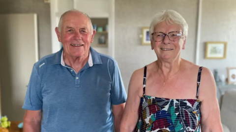 A man and a woman stand side by side in a living room.