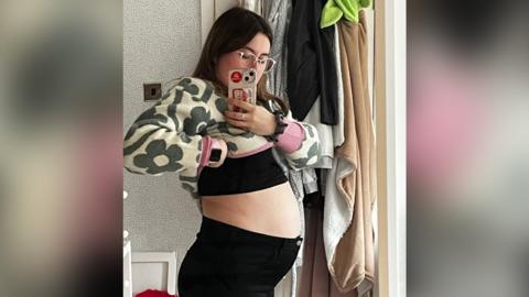 Selfie photo of Natasha Reynolds showing her very bloated stomach. She is wearing exercise trousers, a crop top and glasses.