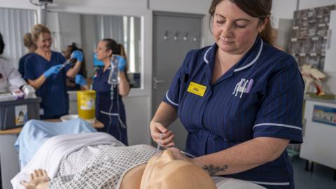 A nurse in blue uniform attaching tubes to a clinical dummy with other nurses holding equipment in the background