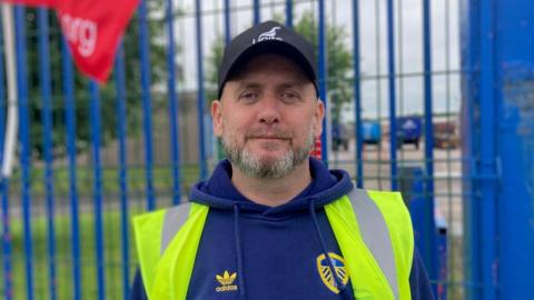 A man wearing a black cap with a white 'Unite' logo. He is also wearing a blue hoodie with a Leeds United logo, and a high-vis jacket. He is standing in front of a blue gate. The man has a grey beard.