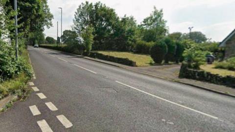A quiet residential road in a suburban or semi-rural area on a bright day. The road runs diagonally from the bottom left corner toward the upper left, gently rising uphill. The surface of the road is dark grey tarmac with white painted markings, including dashed lines along the edge and a solid white line closer to the centre.