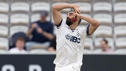 Zafar Gohar about to release the ball as he bowls for Middlesex, with white seats and a few spectators in the background