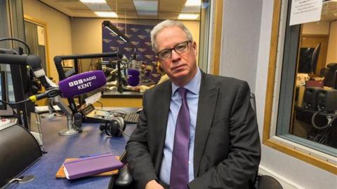 Doug Bannister sits in a chair in the BBC Radio Kent studio with a branded microphone pointing towards him. He is looking at the camera, wearing glasses and a suit with a blue shirt and purple tie