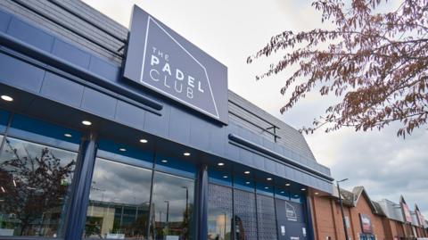 The exterior of the new padel club in Gloucester with branding over a glass entrance, with "The Padel Club" in white letters on a dark blue background