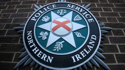 A close-up of a Police Service of Northern Ireland crest on the brown-bricked wall of its Belfast headquarters.  The centre of the crest shows a red cross on a white background, which is inside a white star, inside a green circle, which is inside a larger black circle, all surrounded by silver spikes.  There are six symbols in between the points of the central star, including a harp; a crown; a shamrock; a flaming torch; the scales of justice and an olive branch. 