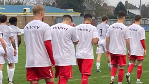 A group of football players, seen from behind, walk in a line on to a green football pitch. They are dressed in red kit with a white t-shirt over the top with the word "Hitchy" printed on the back.