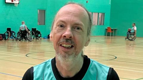 A man with short grey hair inside the sports hall, which has green walls. In the background are wheelchair basketball players using the court.