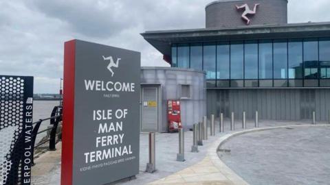 The outside of the ferry terminal, which has large glass windows in the second floor and a three-legs-of Mann symbol above. The is a grey sign that says Welcome - Isle of Man Ferry Terminal in English and Manx to the left and paving in front of it.