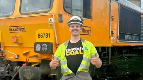 Harry Atkinson in safety hat and overalls, smiling and giving a double thumbs up in front of a coal train