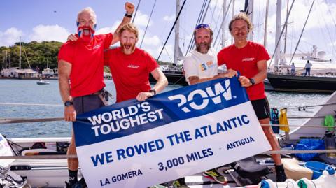 Four men smiling holding a sign which says "World's toughest Row. We rowed the Atlantic. La Gomera - 3,000 miles - Antigua".