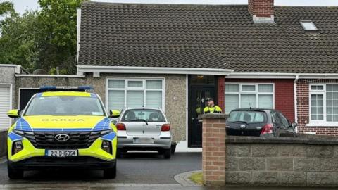 A police officer is seen standing outside the door of a house where two cars are parked along with a police jeep.