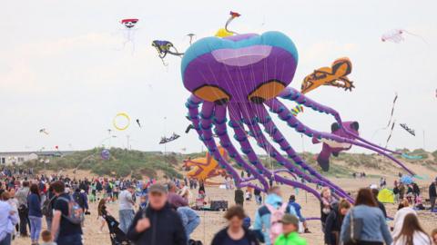 A large purple kite in the shape of an octopus being flown at Redcar's beach. It has large orange eyes. A number of other kites can be seen in the air in the background including an orange lizard. Dozens of people are watching on.